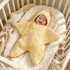 May include: A pale yellow crocheted star-shaped swaddle blanket with a pointed hood and two round buttons on the front. The swaddle features a textured knit pattern and is designed for infants. The text "Photo Prop | Not for sleep" is visible.