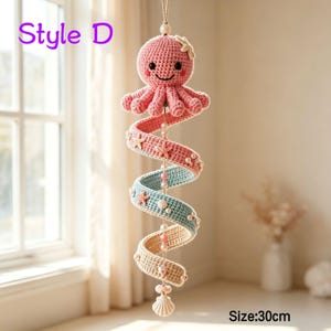 May include: A hanging crochet mobile featuring a pink octopus with a starfish on its head, attached to a spiral ribbon in shades of pink, blue, and beige. The mobile is decorated with small white beads and starfish accents, measuring 30 cm in length.