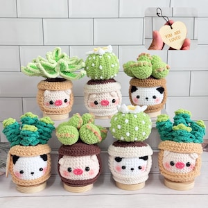 Puede incluir: Colección de decoraciones de plantas de cactus y suculentas hechas a mano con ganchillo. Cada planta está en una maceta decorativa con una linda cara de animal. Las plantas son verdes y blancas, y las macetas son de color beige, marrón y blanco. También se ve un corazón de madera con la frase "YOU ARE LOVED".
