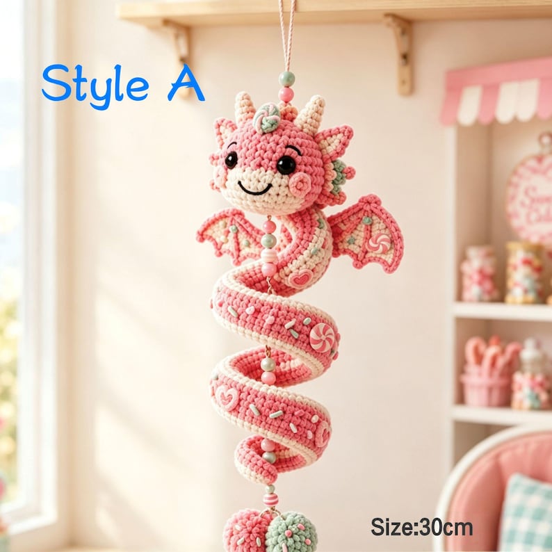 K&ouml;nnte beinhalten: Ein h&auml;ngendes H&auml;kel-Drachenornament in Rosa und Creme mit spiralf&ouml;rmigem K&ouml;rper, kleinen Fl&uuml;geln, schwarzen Augen und bonbonartigen Verzierungen. Der Text Style A und Size:30cm ist sichtbar. Der Drache h&auml;ngt an einer Schnur mit Perlen.