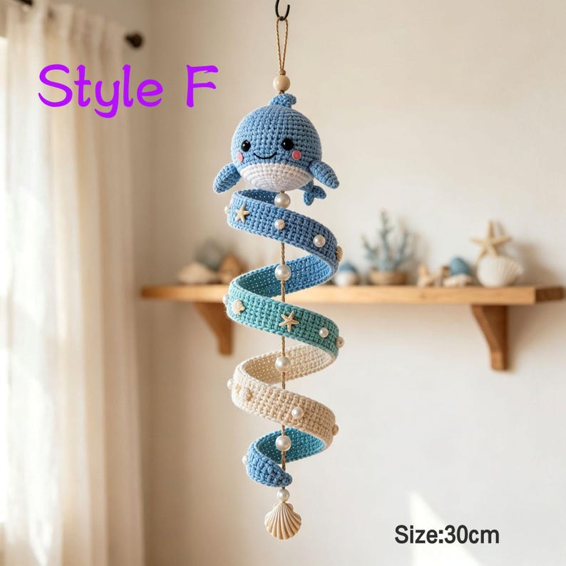 May include: A hanging crochet mobile featuring a blue whale at the top, followed by a spiral strip transitioning from blue to teal to cream, decorated with small white pearls and starfish accents, ending with a seashell. Text reads Style F and Size: 30cm.