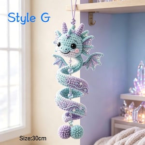 K&ouml;nnte beinhalten: Ein h&auml;ngendes H&auml;kel-Drachenornament mit der Aufschrift Style G und einer L&auml;nge von 30 cm. Der Drache hat einen spiralf&ouml;rmigen K&ouml;rper aus hellblauem und lavendelfarbenem Garn, verziert mit kleinen sternf&ouml;rmigen Details, Perlen und zwei Bommeln.