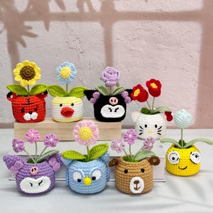 以下が含まれることがあります： 手作りのかぎ針編みの植木鉢のコレクション。それぞれが異なる動物の顔としてデザインされています。鉢にはカラフルなかぎ針編みの花が詰められています。動物には、トラ、アヒル、豚、猫、フクロウ、クマ、そして笑顔の黄色い顔が含まれます。