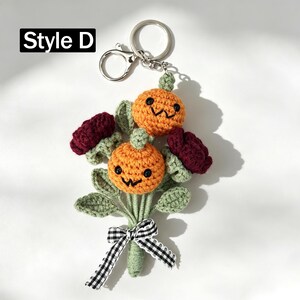 May include: A crochet keychain labeled Style D featuring two orange pumpkins with embroidered black faces, two dark red flowers, and green leaves. The stems are tied together with a black and white gingham ribbon attached to a silver metal ring.