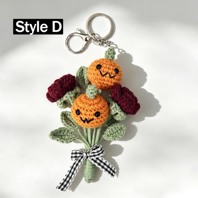 May include: A crochet keychain labeled Style D featuring two orange pumpkins with embroidered black faces, two dark red flowers, and green leaves. The stems are tied together with a black and white gingham ribbon attached to a silver metal ring.