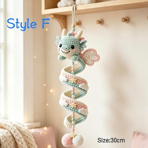 K&ouml;nnte beinhalten: Ein h&auml;ngendes H&auml;kel-Drachen-Mobile mit der Aufschrift Style F, 30 cm lang. Der mintgr&uuml;ne Drache hat rosa Fl&uuml;gel, einen spiralf&ouml;rmigen K&ouml;rper mit Pastellstreifen, kleine wei&szlig;e Wolken, gelbe Sterne und eine Perlenschnur, die in zwei Pompons endet.