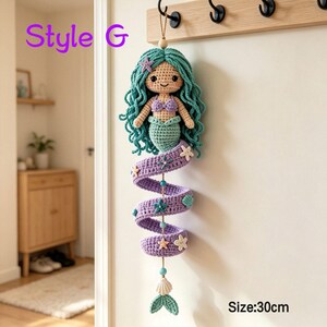 May include: A crochet mermaid wall hanging with teal hair, a purple top, and a spiraling purple tail adorned with small starfish and seashell accents. The figure features a beige face, black eyes, and a smile. Text reads Style G and Size: 30cm.