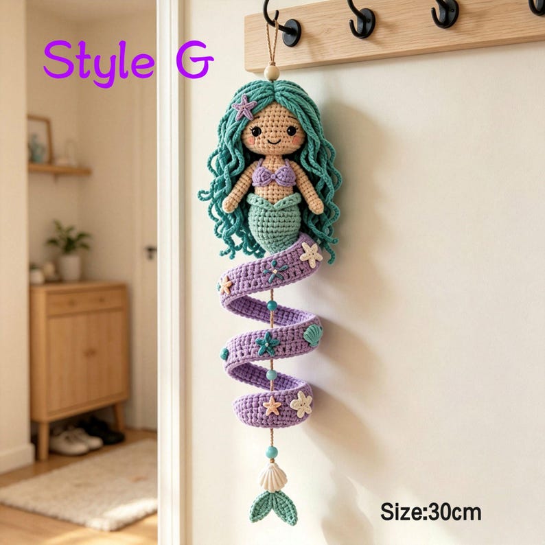 May include: A crochet mermaid wall hanging with teal hair, a purple top, and a spiraling purple tail adorned with small starfish and seashell accents. The figure features a beige face, black eyes, and a smile. Text reads Style G and Size: 30cm.
