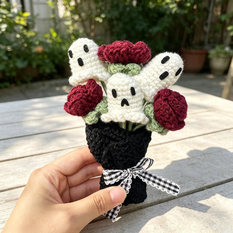 May include: A crochet bouquet featuring three white ghost figures with black embroidered eyes and mouths, interspersed with dark red roses and light green leaves. The stems are wrapped in black yarn and tied with a black and white gingham ribbon.