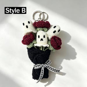 May include: A crochet keychain featuring a bouquet of three white ghost figures with black embroidered faces and three dark red roses with green stems, all wrapped in black yarn with a black and white gingham ribbon and a metal ring. Text: Style B.