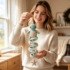 May include: A crochet hanging mobile featuring a light green turtle with a cream-colored shell at the top. Below the turtle, a spiral-shaped crochet strand in light green is decorated with small white beads, tiny seashells, and a cream-colored starfish.