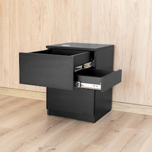 Pode incluir: Uma mesa de cabeceira preta com duas gavetas, ambas parcialmente abertas. A mesa de cabeceira está colocada contra uma parede com painéis de madeira clara e assenta num chão de madeira clara. As gavetas têm ferragens prateadas.