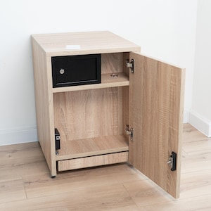 May include: A light oak wood cabinet featuring a single hinged door that is open. Inside, there is a black metal security safe box mounted on the upper shelf, a lower storage compartment, and a small black electronic component on the side wall.