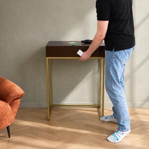 May include: A dark brown console table with a rectangular top and a gold-toned metal frame. The table features a single drawer with a gold handle, standing on four thin metal legs. The design is clean and functional, providing a surface for small items.