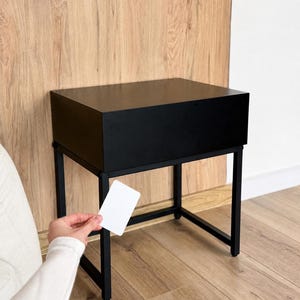 May include: A black bedside table with a rectangular top and a black metal frame. The table is set against a light brown wooden wall and floor. A person's hand holds a white card near the table.
