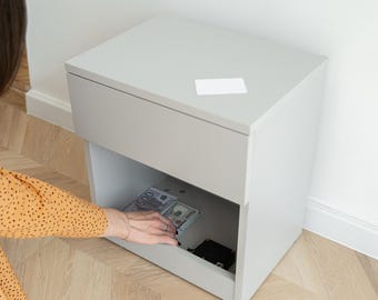May include: A light gray bedside table with a single top drawer and an open storage compartment underneath. A small white rectangular card rests on the flat top surface, while stacks of paper currency are visible inside the open lower shelf.