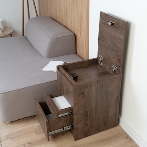 May include: A dark brown wooden bedside table with two drawers and a top compartment with a hinged lid. The drawers are partially open, revealing white interiors. The table is next to a gray sofa.