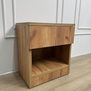 Puede incluir: Una mesita de noche de madera con un cajón y un estante abierto. La mesita de noche está hecha de madera con un patrón de vetas natural y un color marrón cálido. El cajón está situado en la parte superior y el estante abierto debajo.