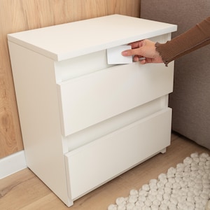 Puede incluir: Una mesita de noche blanca de tres cajones con un diseño rectangular. Una mano saca una pequeña tarjeta blanca del cajón superior. La mesita de noche está colocada junto a una pared de color claro y un sofá gris.