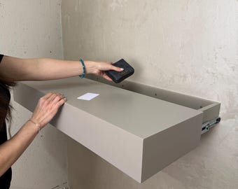 May include: A light gray floating drawer is mounted on a wall. A black wallet with the word "GUESS" is held above the drawer. A white card is on the drawer's surface. The drawer is open, revealing the drawer slides.