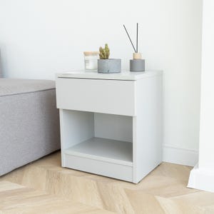 May include: A light gray bedside table with a drawer and open shelf. A small potted cactus, a candle in a glass jar, and a gray diffuser with black reeds sit on top. The table is next to a gray upholstered headboard.