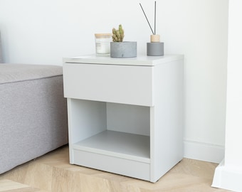 May include: A light gray bedside table with a drawer and open shelf. A small potted cactus, a candle in a glass jar, and a gray diffuser with black reeds sit on top. The table is on a light wood floor.