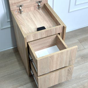 May include: A light oak wood-grain bedside table featuring a top compartment with a hinged lid and two pull-out drawers below. The unit has metal hinges and drawer slides, with a white interior lining visible inside the open top drawer.