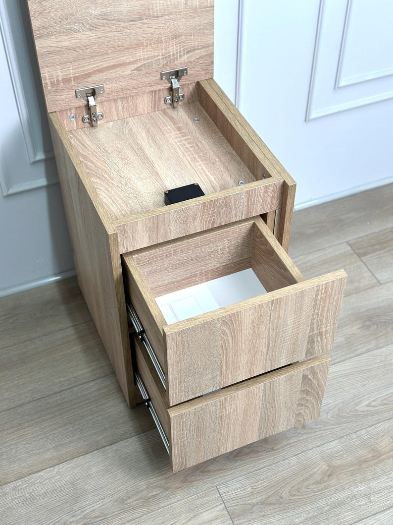 May include: A light oak wood-grain bedside table featuring a top compartment with a hinged lid and two pull-out drawers below. The unit has metal hinges and drawer slides, with a white interior lining visible inside the open top drawer.