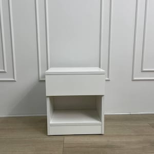 May include: A white nightstand with a single drawer and an open shelf below. The nightstand is rectangular and sits on a light brown wooden floor. The background is a white wall with vertical molding.