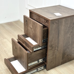 May include: A dark brown wood-grain storage unit featuring three tiered, pull-out drawers with visible metal sliding tracks. The drawers are stepped in a staircase arrangement, each revealing a white interior base and a flat top surface.