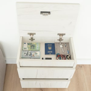 May include: A white wooden nightstand with an open top drawer revealing cash, a passport, and jewelry. The nightstand has two additional drawers below and is set against a white wall and light wood floor.
