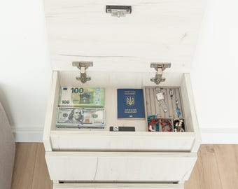 May include: A white wooden bedside cabinet with an open top drawer containing banknotes, a passport, and jewellery. The cabinet has two further drawers below and is against a white wall and light wood floor.