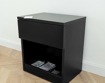 May include: A matte black nightstand featuring a flat top surface with a small white rectangular card, a single upper drawer, and an open lower storage compartment containing stacks of paper currency including US dollar and Euro banknotes.