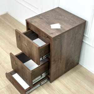May include: A dark brown wood-grain finish mobile storage unit featuring three open drawers with white interiors and silver metal sliding tracks. The top surface is flat and rectangular, and the unit is constructed from composite wood panels.