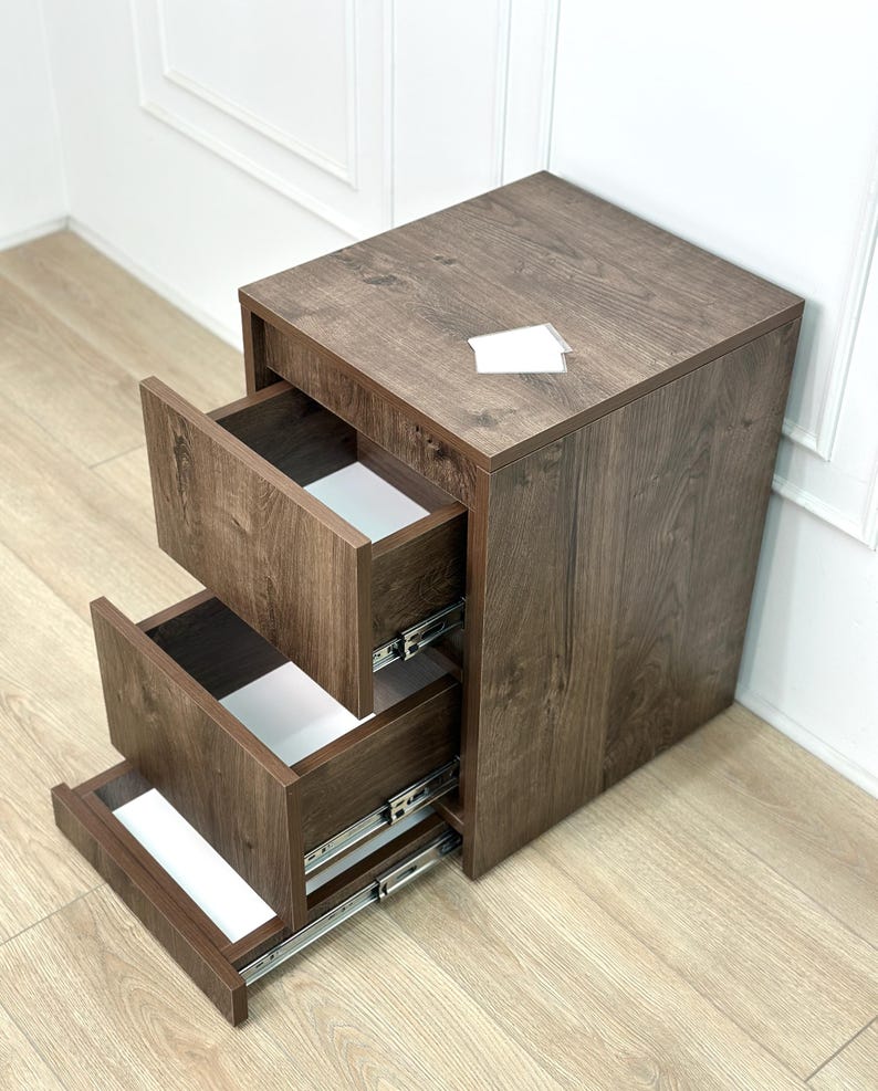 May include: A dark brown wood-grain finish mobile storage unit featuring three open drawers with white interiors and silver metal sliding tracks. The top surface is flat and rectangular, and the unit is constructed from composite wood panels.