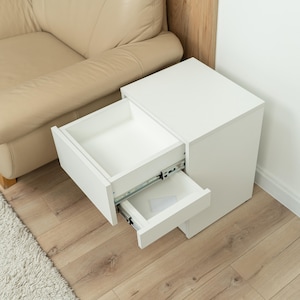 Könnte beinhalten: Ein weißer Nachttisch mit zwei offenen Schubladen. Die Schubladen befinden sich auf Metallschienen und geben den Innenraum frei. Der Nachttisch steht neben einem beigen Sofa und auf einem hellen Holzboden.