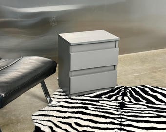 Può includere: Un comodino grigio chiaro dotato di due cassetti sovrapposti con una finitura liscia e opaca. L'unità rettangolare presenta una superficie superiore piatta e una silhouette pulita e squadrata, offrendo una soluzione di archiviazione.