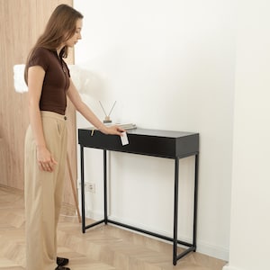 Puede incluir: Una consola negra con una tapa rectangular y un marco de metal minimalista. La mesa está colocada contra una pared blanca. La mesa mide 122 cm de largo, 30 cm de ancho y 76 cm de alto.