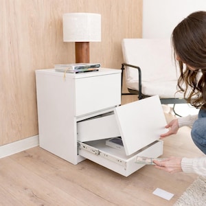 Pode incluir: Uma mesa de cabeceira branca com duas gavetas, uma parcialmente aberta. Uma lâmpada com uma sombra branca está no topo, juntamente com uma pilha de livros. A mesa de cabeceira está numa sala com piso de madeira clara e uma cadeira.