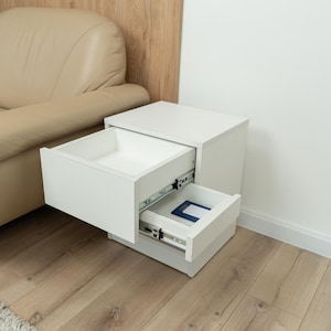 May include: A white bedside table with two open drawers featuring metal sliding tracks. The top drawer is empty, while the bottom drawer contains a small blue and white rectangular object. The unit has a smooth, matte finish and a square design.