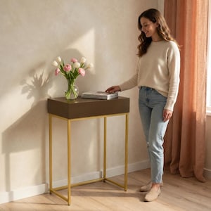 May include: A brown rectangular side table with gold-colored legs. A clear vase with pink and white flowers sits on top of the table, along with a stack of books. The table is in a room with a woman standing next to it.