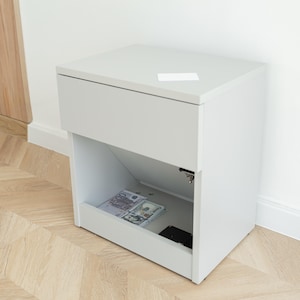May include: A white bedside cabinet with a drawer and an open compartment. The open compartment reveals stacks of cash and a black object. A white rectangular object rests on the cabinet's top.