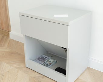 May include: A light gray bedside table featuring a flat top surface with a small white rectangular card, a single upper drawer, and an open lower storage compartment containing stacks of paper currency and a small black electronic device.