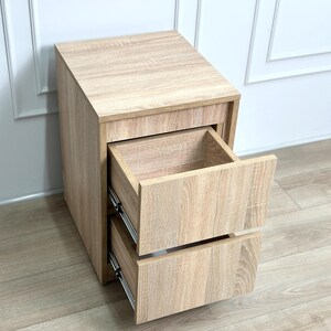 May include: A light oak wood grain bedside table featuring two open drawers with metal side glides. The rectangular unit has a flat top surface and a vertical orientation, showcasing a consistent light brown wood texture throughout the entire structure.