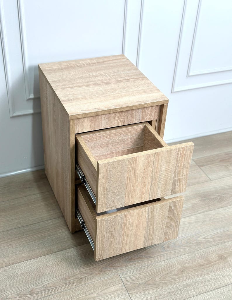 May include: A light oak wood grain bedside table featuring two open drawers with metal side glides. The rectangular unit has a flat top surface and a vertical orientation, showcasing a consistent light brown wood texture throughout the entire structure.