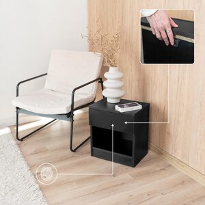 Könnte beinhalten: Ein schwarzer Nachttisch mit Schublade und offenem Regal, neben einem weißen Stuhl mit schwarzem Metallrahmen. Eine weiße Vase mit getrockneten Zweigen steht auf dem Nachttisch, zusammen mit einem Smartphone. Der Nachttisch steht auf einem hellen Holzboden.
