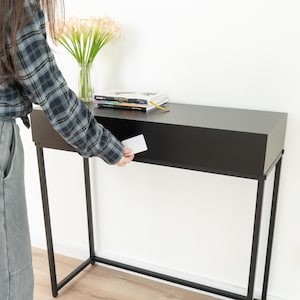 May include: A black console table with a slim rectangular top and a thin metal frame. The table features a hidden drawer compartment being opened with a white card. A glass vase with light pink flowers and a stack of books sit on the surface.