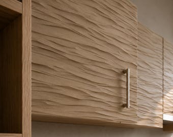 May include: A light brown wooden cabinet door featuring a textured, wavy, horizontal relief pattern across its surface. The cabinet door is equipped with a single, vertical, cylindrical metal handle in a brushed silver finish.