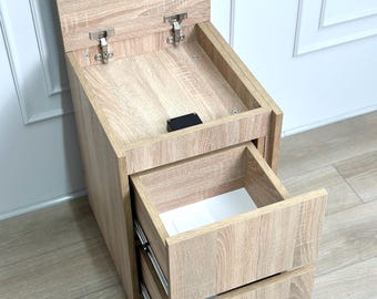 Può includere: Un mobile contenitore con finitura effetto legno di rovere chiaro, dal design verticale con coperchio incernierato superiormente che rivela uno scomparto e due cassetti inferiori estraibili con guide metalliche e un fermo nero.