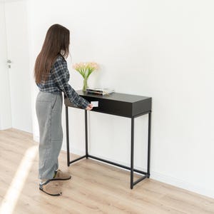 Könnte beinhalten: Ein schwarzer Konsolentisch mit rechteckiger Platte und Schublade. Der Tisch hat ein minimalistisches Design mit dünnen, schwarzen Metallbeinen. Eine Vase mit rosa Blumen und einige Bücher stehen auf dem Tisch.